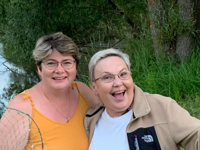 Two women are pictured in an outdoor setting, with lush green foliage and a body of water visible in the background. The woman on the left, wearing a yellow tank top, has short, light brown hair and is smiling. She is holding a fishing net. The woman on the right, with short, white hair, wears glasses and a light brown jacket over a white t-shirt. She is also smiling broadly. The scene suggests a relaxed, possibly recreational moment. The location is identified as Suravatíkha, Russia.