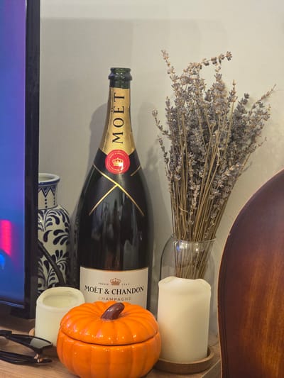 The image depicts a still life arrangement on a wooden surface, likely a shelf or table. Dominating the center is a large bottle of Moët & Chandon champagne with its signature gold foil and red seal visible. To the right of the champagne bottle, a clear glass vase holds a dried bouquet of lavender. In the foreground, a bright orange pumpkin-shaped lidded container sits between two white pillar candles, one to its left and another to its right. To the far left of the frame, partially visible, is the edge of a black television screen displaying a blurred purple and pink glow. Behind the objects, a light gray wall serves as the backdrop. The arrangement suggests a festive or decorative setting. No people are present, and no specific location in Kâğıthane, Turkey, can be identified from the visual information.