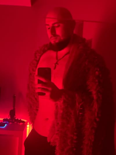 A person, illuminated by red light, stands in front of a mirror, taking a selfie with a smartphone. The individual is wearing a fuzzy, dark-colored coat and a necklace with a cross pendant. They have a shaved head and facial hair. The background appears to be an indoor environment with red walls. To the left, a white table is visible, with some items on it, including a device with blue lighting. The scene is likely a private residence. There is no discernible connection to the city of Gliwice, Poland, nor are there any visual cues that would suggest this specific location.