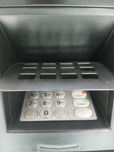 A close-up view of an ATM's pin pad. It features a standard numerical keypad layout with numbers 0-9, "Cancel" and "Enter" buttons. The "Cancel" button is red, and the "Enter" button is green. A protective shield with rectangular cutouts covers the top portion of the keypad. The buttons appear well-used, showing wear. The surrounding structure is black, made of plastic.