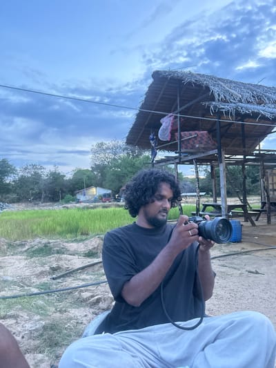 A man with curly dark hair is holding a black camera with a large lens to his face, as if taking a photo. He is wearing a dark t-shirt. His right hand is on the camera, and his left hand is around the lens. A thin silver band is visible on his ring finger. Next to him, a person in light-colored pants sits with their legs crossed. They are positioned to the right of the man with the camera.

The background shows a rustic, open-sided structure with a thatched roof. Inside the structure, there are some visible objects, including what appears to be a red and black checkered mat or blanket, and a pink net. Beyond the structure, a green field, possibly a paddy field, stretches out. In the distance, there are trees and a small building with a yellow light emanating from a window.

The sky is cloudy and appears to be a pale blue, suggesting it might be late afternoon or early evening. The overall scene has a natural, outdoor feel. The man's focus on the camera suggests he is actively engaged in photography. The location, Shastrawela, Sri Lanka, provides a geographical context for the rural setting with the paddy field and rustic structures. No text is visible in the image.