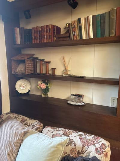 This interior scene depicts a dark wood built-in shelving unit above a seating area, located in Monterey, United States of America.

The top shelf contains approximately 30 books, varying in size and color. A collection of reddish-brown bound books is centrally located, flanked by other books with dark and light-colored spines, one titled "American Natural History." A gold-colored duck figurine stands on the right, functioning as a bookend, with two dark-spined books stacked horizontally beneath it. Two black wall-mounted spotlights are visible above this shelf.

The middle shelf features a wicker basket with a hinged lid, containing a patterned cushion. To its right are four wooden spools or bobbins, graduated in height, with the tallest exhibiting a green-painted top edge. Further right, a clear glass reed diffuser bottle with reeds and a small, dark, ornate tray are present.

The bottom shelf displays a decorative plate with a floral pattern, propped vertically on a dark stand. Next to it, a clear glass vase holds a bouquet of artificial pink and green roses. On the right, a silver-colored oval tray contains two clear, patterned drinking glasses and a stack of dark coasters. A white wall-mounted panel with "PHONE" and "TV" labels and three small icons is visible on the far right.

Below the shelving unit, the top portion of a seating arrangement, likely a sofa or bench, is visible. It is upholstered in a floral pattern featuring pink, green, and brown tones. Multiple throw pillows are present, including one with a light brown houndstooth pattern and another in a solid light blue color. A dark fabric is partially visible beneath the pillows. The wall behind the shelves is white with vertical paneling. No individuals are depicted.