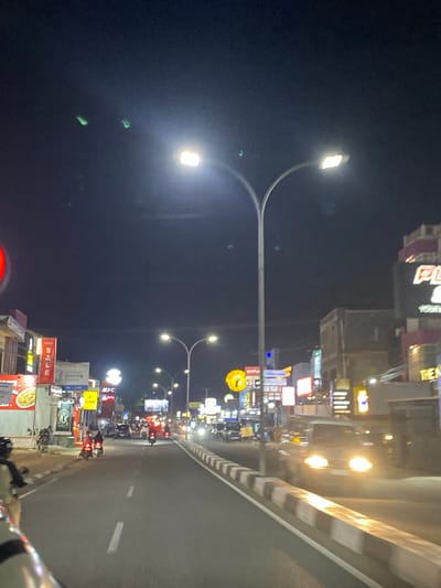 This media file captures a nighttime scene on a street in Watampone, Indonesia. The perspective suggests the image was taken from a moving vehicle, likely a motorcycle, as indicated by the blurred foreground elements.

The primary subjects are the street and the buildings lining it, illuminated by various light sources. There are streetlights with multiple lamps, casting a bright glow on the dark sky. The buildings are adorned with numerous illuminated signs displaying advertisements and information, some of which are legible. Visible text includes "KFC", "SALE", and what appears to be "Learning Centre". Other signs have graphics and text in an unknown language, but the overall impression is that of a commercial or entertainment district.

Several vehicles, predominantly motorcycles with riders, are visible on the road, suggesting traffic flow. The activity taking place is ordinary urban life at night, with people commuting or perhaps engaged in leisure activities. The time of day is clearly night, with a dark sky and artificial lighting dominating the scene. The weather appears clear, with no indications of rain or fog. The mood conveyed is one of a bustling, vibrant urban environment at night. The perspective and the presence of traffic suggest a dynamic, lively atmosphere.