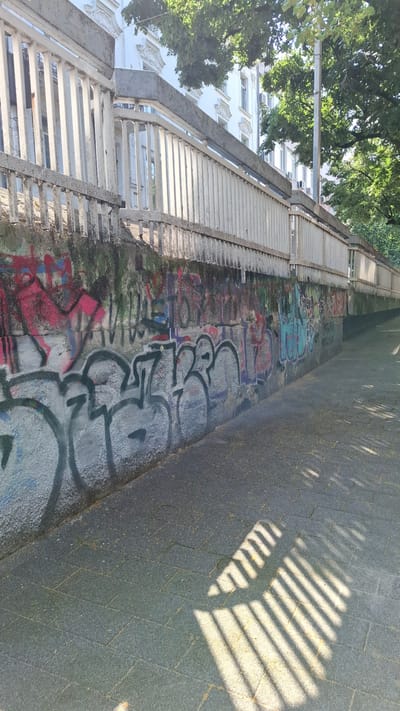 The image depicts a graffiti-covered concrete wall along a paved walkway. The wall is topped with a white railing and fence, beyond which a building with white architectural details and trees are visible. The walkway is paved with rectangular stones and is partially covered in a light scattering of yellow debris. Sunlight casts striped shadows from the railing onto the walkway. The graffiti on the wall is multicolored and includes both large, bold lettering and more abstract designs. The overall scene suggests an urban environment, likely a public thoroughfare or underpass in Plovdiv, Bulgaria, given the context provided. There are no people or animals present in the image.