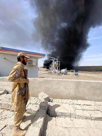 An outdoor scene in Lundey Kalay, Afghanistan, depicts a uniformed individual observing a large column of black smoke. In the left foreground, a male with a beard and a tan cap stands angled to the left, gazing towards the right. He wears a tan camouflage uniform and tan boots. His right hand holds an AK-pattern rifle, which is pointed downwards. Behind him and to his left is a white building with blue and red trim on its roofline and a "GMCC" branded air conditioning unit mounted on its exterior wall. The immediate foreground consists of broken concrete blocks and rubble on a dirt surface. A concrete barrier wall separates the foreground from the middle ground. In the middle ground, beyond the wall, are industrial elements including a white and yellow metal structure with blue piping, a horizontal grey cylindrical tank, and a blue drum lying on its side, all situated on a dirt or gravel area. Dominating the right half of the image and the background is a large, dense plume of black smoke rising vertically into a partly cloudy sky, indicating a significant fire source.