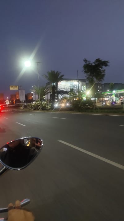 The image captures a scene at dusk on a street in Pusakarakyat, Indonesia. A street light with a bright, blown-out glow illuminates the left side of the frame. In the background, a multi-story building with various signage, including "1M" and "25", is visible. A green canopy, likely part of a gas station, is also present. Palm trees and other foliage are scattered throughout the scene. A motorcycle's side mirror is prominently featured in the foreground, reflecting the sky and the rider's upper body and hand. The road itself has white lane markings. The overall atmosphere suggests a busy urban environment transitioning from day to night.