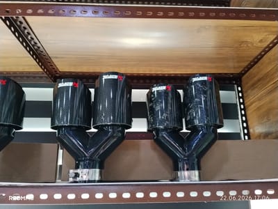 The image shows a close-up view of four carbon fiber exhaust tips with the Akrapovič logo, displayed on a metal shelf unit. The exhaust tips are black with a checkered pattern, and each has a red Akrapovič logo. They are arranged in two pairs, with each pair connected by a Y-shaped pipe. The background consists of wooden shelves and metal framing. The lighting suggests an indoor setting, possibly a store or workshop. Visible text on the bottom of the image includes "REDMI 95" and a date and time stamp "22.06.2026 17:06". The location is Yovon, Tajikistan.