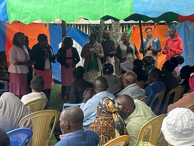 The image depicts an outdoor community gathering taking place under a large, colorful canopy made of green, blue, and orange tarpaulins in Kiganjo, Kenya.

In the foreground, a group of approximately ten individuals, predominantly women, are standing. Several women on the left side are dressed in similar pink skirts paired with white or dark tops, and some hold microphones, indicating they are either performing, singing, or addressing the audience. On the right, another set of individuals, including women and one man in a red hoodie, also stand under the canopy, possibly part of the same group or facilitating the event.

Facing the standing group is a diverse audience of men and women, seated on rows of light yellow and blue plastic chairs. Most audience members have their backs or sides to the camera, appearing attentive and engaged in the proceedings. The ground beneath them is natural, likely grass or dirt.

The activity appears to be a public or community event, potentially a church service, a local performance, or a meeting, given the setup with performers/speakers and an audience. The time of day is daytime, with natural light illuminating the scene, though the canopy provides shade. The visible text "HTENG WEAR" is printed on the back of a grey sweatshirt worn by a male audience member. The overall mood appears communal and orderly.
