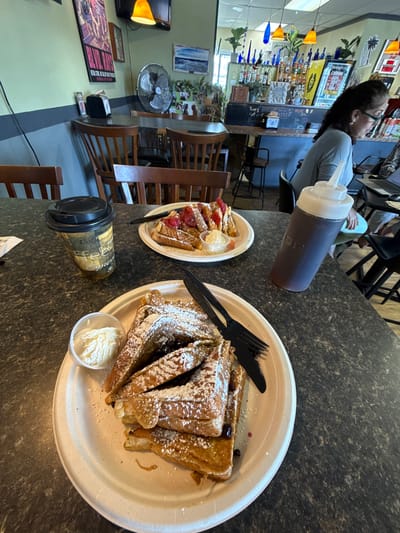 This media file captures an indoor scene within a casual cafe or diner, likely during daylight hours given the breakfast items. The primary focus is a table laden with food and drinks.
In the foreground, two plates of French toast are visible on a dark, speckled countertop. The larger plate, on the right, holds multiple pieces of French toast generously dusted with powdered sugar and garnished with blueberries. It's accompanied by a small plastic cup of whipped cream or ice cream and a black plastic fork and knife. The smaller plate, on the left, features French toast topped with sliced strawberries and powdered sugar, also with a small cup of what appears to be a sauce or whipped cream. Between the plates, a coffee cup with a lid and a brown-gold design (possibly indicating "Premium Roast Coffee") rests on the table. A dark-colored squeeze bottle, likely containing syrup, is positioned further back on the table.
In the mid-ground and background, the setting becomes clearer. A person with dark, wavy hair and glasses, wearing a light blue top, is seated at a counter or bar to the left, facing towards the right side of the frame. Their profile is visible. Another person's legs are partially seen behind them. The bar area behind the person is well-stocked with various bottles, and decorative pendant lights (orange and blue) hang above. A chalkboard behind the bar shows partial text, including "LATTE'S." The walls are adorned with framed pictures and art. To the far left, a screen or television is visible, and on the floor, a large poster reads, "THE WORLD WILL BE ON IT'S KNEES." A ceiling fan and wooden chairs are also part of the room's furnishings.
The image suggests a relaxed meal at a local eatery in Hampton, United States of America.