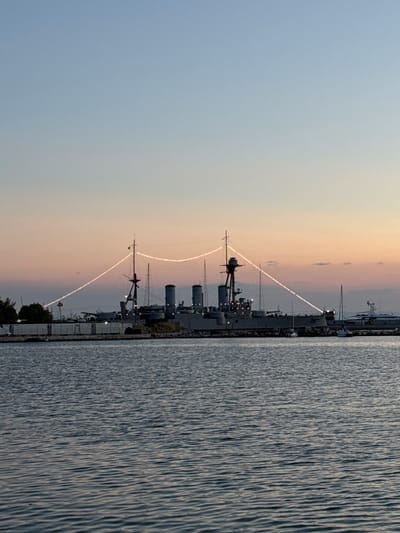This media file depicts a twilight scene with several large, gray ships docked along a waterfront. The sky transitions from a pale blue at the top to a soft orange and pink hue near the horizon, indicating it is likely sunset or sunrise. The water in the foreground is choppy, with visible ripples and reflections of the ships and sky.

The ships appear to be historical vessels, possibly warships, with prominent smokestacks and mast-like structures. One ship in particular has a series of lights or reflective elements arranged in a line that extends from its upper structure upwards and across, creating a distinctive illuminated trail against the sky. The overall atmosphere is serene and tranquil, with the fading light casting a peaceful glow over the scene. The location is identified as the Municipal Unit of Dafni, Greece, suggesting a harbor or naval museum setting. There is no visible text in the image.