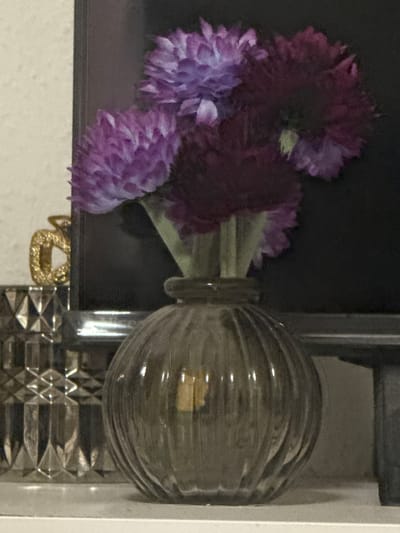This image, captured in Vellmar, Germany, depicts a still life arrangement of decorative objects on a light-colored, flat surface. In the foreground-center, a globular, vertically ribbed, translucent dark gray glass vase holds a bouquet of five artificial flowers. Three of the flowers are light purple-mauve, while two are dark plum-burgundy, all featuring dense, textured petals. Their light green stems are visible within the vase's narrow neck.
To the left of the vase, a clear glass object with a faceted, diamond-cut pattern is partially visible. Behind this, a glittery gold-toned decorative object, resembling a stylized letter 'C' or similar curvature, is discernible.
The immediate background behind the vase and flowers is a dark, featureless vertical surface, potentially a television screen or dark furniture, showing faint reflections. To the left of this dark surface, a light-colored wall with a subtle textured finish is present. No individuals or active interactions are depicted.