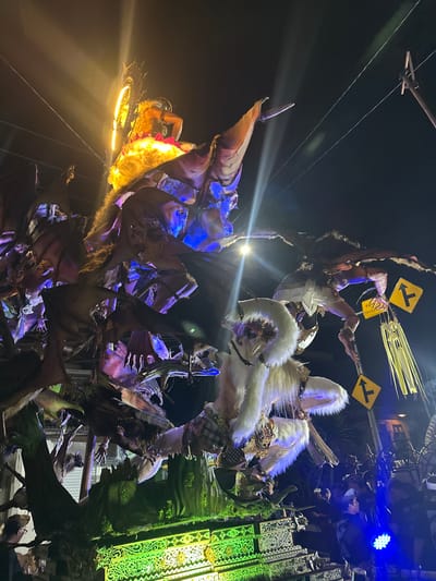 This nighttime image depicts a large, intricately constructed parade float, likely an Ogoh-Ogoh, illuminated with various lights, in Munggu, Indonesia. The float features multiple grotesque and mythical figures. At the top, a humanoid figure with a reddish-orange lower body glows intensely, framed by a bright yellow halo-like structure. Below it, several dark, bat-like or winged creatures with visible limbs and faces are intertwined among tree-like structures, illuminated by blue spotlights. In the foreground, a prominent white, furry creature with a mask-like face, dressed in traditional Balinese checkered cloth (poleng), crouches on the float. To its right, another creature or figure holds two yellow, diamond-shaped road signs depicting "T" intersection symbols, with one also bearing Indonesian text "Kidi` cecepa...". The base of the float is elaborately carved and illuminated with vibrant green lights. Overhead power lines are visible against the dark night sky. A crowd of people, some wearing traditional attire, is visible at the bottom right, observing the procession.