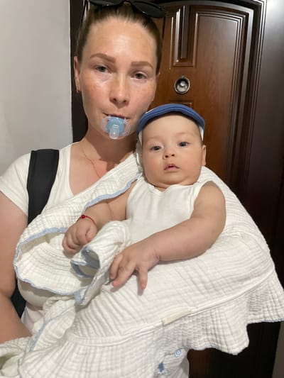 The image shows a woman holding a baby. The baby is wearing a white onesie and a blue headband. The woman is wearing a white tank top and a black strap across her chest. A blue pacifier is in the woman's mouth, and the baby is wrapped in a white blanket with blue trim. The background appears to be indoors, with a dark wooden door visible behind them. The lighting suggests it could be daytime. The overall scene is intimate, with the woman cradling the baby close to her. No visible text is present in the image.