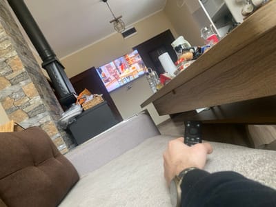 The image depicts an indoor scene in Kozarsko, Bulgaria, observed from a first-person perspective. In the immediate foreground, a person's right hand, wearing a wristwatch on the left wrist and a dark long-sleeved garment, holds a black remote control. The hand rests on a light-colored, possibly fabric-covered, surface, likely a sofa or armchair, with a brown upholstered section visible to the left.
In the midground, a dark wooden coffee table or similar furniture piece is positioned, its surface cluttered with various items including a white roll of paper towels, a clear pitcher containing red liquid or an object, multiple packaged snack items, and a silver multi-tiered container. The floor beneath the table appears dark.
The background reveals a living space. A large flat-screen television is mounted on a light beige wall, displaying an indoor scene with multiple figures, possibly from a show or movie. To the left of the television, a black freestanding wood-burning stove with a tall black flue pipe is situated against a wall accented with multi-toned stacked stone veneer. A dark wooden door frame is visible to the left of the TV. The ceiling features a decorative pendant light fixture and a rectangular white vent.