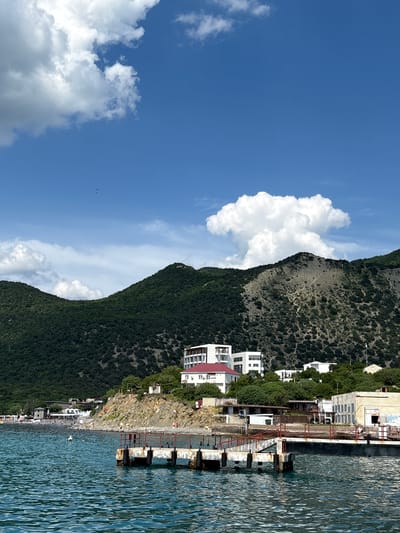 This image depicts a coastal scene in Bol'shoi Utrish, Russia, featuring a body of water in the foreground, a shoreline with structures, and two mountains in the background under a blue sky.

The sky occupies the upper portion, displaying a vibrant blue with several white cumulus clouds. A particularly large, distinct, mushroom-shaped cumulus cloud is prominent above the right mountain. A small, dark, winged object, possibly a bird or a distant small aircraft, is visible high in the sky to the left of the image center.

Two heavily forested mountains dominate the mid-ground and background. The left mountain is uniformly green with dense tree cover. The right mountain, also largely green, features a significant, lighter-toned, sparsely vegetated area of exposed rock or scree on its upper slope, creating a visual contrast.

At the base of the mountains, along the shoreline, numerous buildings are present. Several multi-story white buildings, some with red roofs, are situated on the slopes and closer to the water. An older, light-colored, possibly industrial or abandoned building is visible near the water's edge on the right side.

A concrete pier with a rusty red metal railing extends into the blue-green, rippled water from the right side of the shoreline. The pier is supported by concrete pillars. A smaller, broken or incomplete pier structure, also concrete, is positioned in the water slightly to the left and closer to the viewer than the main pier.

The shoreline to the left of the main pier is a rocky beach where several small human figures are discernible, indicating people are present on the beach. One individual, appearing as a white form, is swimming in the water to the left of the main pier's end.