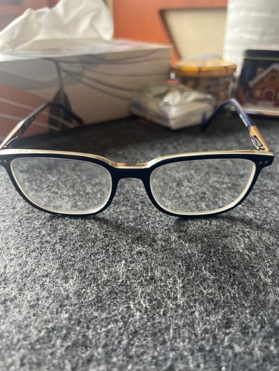 The scene depicts an indoor environment in Ittenheim, France. No individuals are present. In the foreground, a pair of eyeglasses with dark blue frames and gold-colored accents on the top rim and temples rests centrally on a dark gray, fibrous surface. The lenses are clear. The background is out of focus but reveals several objects. To the left, a white tissue box with a pulled tissue is visible. To the right, a clear plastic bag containing small white items, a round amber-colored jar with a gold lid, and a rectangular item displaying a red-roofed house are discernible. The immediate environment appears to be a tabletop.