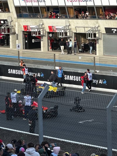 The image depicts a motor racing event at the Circuit de la Sarthe, Le Mans, France, viewed from an elevated spectator area. A chain-link fence partially obscures the immediate foreground, behind which a crowd of people observes the scene.
The midground features a concrete pit lane with white markings. A black barrier wall with prominent "SAMSUNG Galaxy AI" branding separates the pit lane from the pit garages. Multiple individuals, identified as event personnel or team members, are present on the track. At least three open-wheel racing vehicles are visible: a red car, attended by personnel wearing vests numbered "402" and "403"; a black and yellow car displaying "42" on its rear wing; and a black car with the number "97" on its side, facing left. Personnel, some wearing numbered vests (e.g., "30," "374," "396"), are observed walking, standing in groups, and attending to the race cars. A camera on a tripod/dolly is positioned on the track surface. A person in an orange suit is partially visible near the pit wall.
In the background, a multi-level concrete building houses pit garages on the lower level, indicated by numbers "28" and "02." Advertisements for "Erborian Korean Skin Therapy" are visible within the garages. Above the garages, multiple enclosed viewing boxes contain groups of spectators. "Boursorama Bank" and "Hero" branding are displayed prominently on the upper levels of the building. The scene is illuminated by bright daylight, casting distinct shadows.