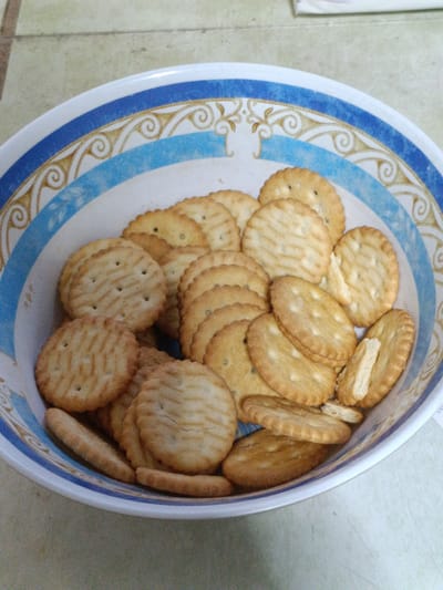 A high-angle close-up shot shows a white bowl with a blue and gold decorative rim filled with round, light brown crackers. The crackers are stacked on top of each other, with some overlapping. A few crackers are broken, and one is standing upright on its edge. The bowl is placed on a light-colored, textured surface. There is no identifiable location in the city of Martuni, Armenia, or any people present in the image.