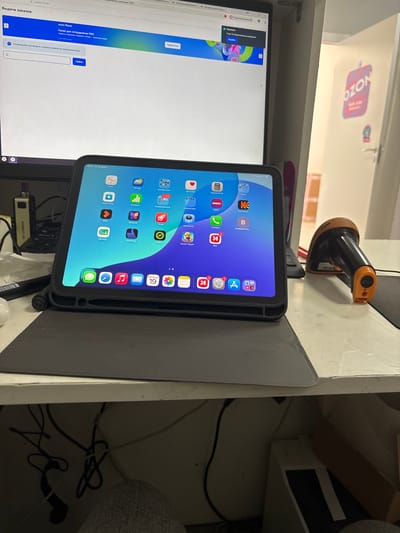 The image shows a workspace with a tablet and a monitor. The tablet is displaying a home screen with various app icons, including "Health", "Calculator", "Phone", and "Apple Store". The monitor is displaying a webpage with Russian text, likely related to order fulfillment or a delivery service, as indicated by phrases like "Выдача заказов" (Order Issuance) and "ozon банк" (Ozon Bank). A barcode scanner is visible mounted on the wall above the tablet. The scene appears to be indoors, possibly an office or a home workspace, with a slightly cluttered background suggesting a functional area. The time indicated on the tablet is "15:51. Пт 19 июня" (15:51. Friday, June 19th), suggesting it is afternoon. A sticker with "NOZO" is visible on the wall in the background. The overall impression is that of a busy work environment where a tablet is being used for tasks related to managing orders or inventory, supported by the presence of a barcode scanner.