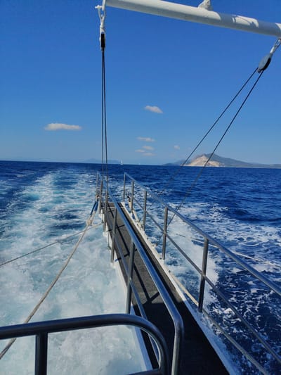 The image shows the wake of a boat sailing on the deep blue sea under a clear blue sky with a few scattered clouds. The boat is moving away from the viewer, with white foam and turbulent water trailing behind. The perspective is from the stern of the boat, looking back. A metal railing and part of the deck are visible in the foreground. In the distance, a mountainous island with a light-colored slope is visible on the horizon, and a few distant sailboats can be seen. The scene appears to be set in the Aegean Sea, possibly near the coast of Kastri, Greece, given the distinctive mountainous landscape and clear blue waters typical of the region. There are no people or animals visible in the frame. The overall impression is one of serene travel on a sunny day at sea.