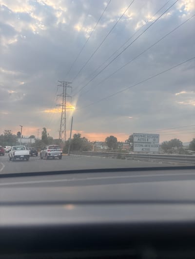 The image captures a hazy, late afternoon or early morning scene on a multi-lane road, likely taken from inside a moving vehicle in Tultitlán, Mexico. The sky is partially cloudy, with the sun breaking through the clouds, casting a warm, golden glow near the horizon and illuminating the atmosphere.

A large, metallic electricity transmission tower stands prominently on the left side of the frame, with numerous power lines extending across the sky. Several vehicles, including a white pickup truck and a silver SUV, are visible on the road, suggesting slow-moving traffic or a queue. A guardrail or barrier runs along the right side of the road, partially obscured by the vehicle's interior window frame.

A white rectangular sign is visible on the right side of the road. The text on the sign, in Spanish, reads: "PREPARE SU CUOTA / AUTOS $ - -.00" (Prepare your toll / Cars $ - -.00), with the exact fee obscured or not filled in. Below this, it reads: "SISTEMA ANTIEVASION ACTIVO" (Anti-evasion system active). This indicates the approach to a toll booth or a section of a toll road. In the background, beyond the road and structures, sparse trees and some distant buildings are visible. The overall mood is calm, emphasized by the soft, diffused sunlight.