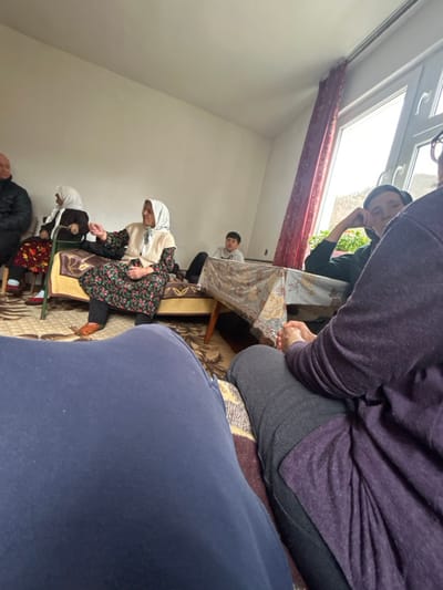 An indoor scene in Erma Reka, Bulgaria, depicts a gathering of at least seven individuals within a modestly furnished room. The walls are light-colored, and a large window on the right features a partially drawn red curtain, revealing an outdoor landscape beyond. The floor is covered with a patterned rug.
From the left, a bald man in a dark jacket is partially visible. Next to him, a woman wearing a white headscarf and dark clothing is seated on a bed or divan, holding what appears to be a green walking stick. Centrally positioned, an elderly woman in a white headscarf, a light-colored vest, and a dark floral dress sits on a bed, gesturing with her right hand. Her feet are visible, clad in orange-brown shoes. Behind a small table covered with a patterned cloth, a young boy in a light grey top is seated. To the right, a man wearing a dark cap is seated on another bed, resting his head on his left hand, looking towards the left.
The foreground is dominated by the legs and laps of two individuals from the photographer's perspective: a lap wearing dark blue casual trousers on the left, and a lap wearing grey trousers and a dark purple top with hands clasped on the right. The room contains two beds or divans with patterned bedspreads and a small wooden table. The individuals appear to be engaged in conversation or social interaction within a residential setting.