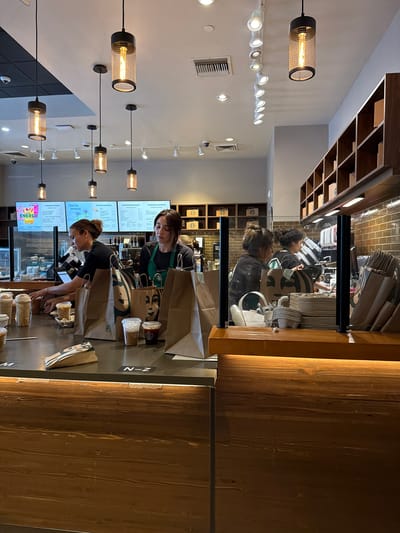 The image depicts the interior of a Starbucks location in Indian Wells, USA, identifiable by the Starbucks employee aprons and menu boards. Two employees are visible behind the counter, seemingly preparing orders. One employee is closer to the camera, reaching down toward an item on the counter, while the second employee is slightly behind and to the right. Several paper bags and cups of various sizes are arranged on the counter, some with the Starbucks logo visible. The foreground of the image is dominated by a wooden wall or partition to the right and the counter with items in front of it. The lighting is a mix of warm pendant lights and cooler recessed lighting in the ceiling. Security cameras are also visible in the ceiling. The scene appears to be a typical service area in the Starbucks, with employees filling orders for customers.