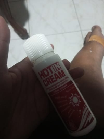A hand holds a bottle of "Hot in Cream" pain relief ointment. The bottle is white with a white cap, and red lettering. The label states it relieves aches, joint pain, and muscle aches, and is non-sticky. The bottle indicates it is for aches, stiff muscles, joint pain, headaches, and bloating. A foot with a bandage on the ankle is visible in the background. The setting appears to be a room with tiled flooring.