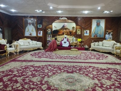 The image shows a large room with ornate wooden walls and a large, intricately patterned carpet covering the floor.  The room is furnished with two white, ornately carved sofas. A single man is seated on a large golden chair, wearing a white robe and a white hat. Another man is kneeling in front of him, wearing a red robe.  The walls are adorned with framed portraits of men in traditional dress,  all framed in ornate gold frames. The room has a number of light fixtures in the ceiling and a decorative archway leading to another room. The scene is likely taking place in the Emir's Palace in Kaltungo, Nigeria.  The coordinates provided place the location in the center of this town.  The image appears to show a traditional, formal ceremony, possibly a meeting between a high-ranking official and a visitor.  The postures of the men and the ornate setting suggest a formal and hierarchical relationship.  The environment is rich and elegant. The overall impression is one of authority, respect, and tradition.
