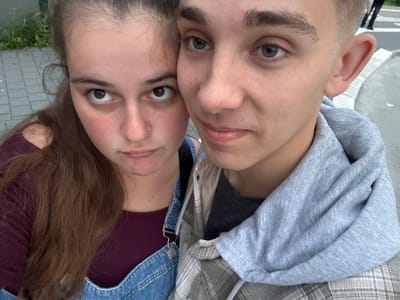 A close-up, outdoor, eye-level selfie shows two young people, a female on the left and a male on the right, positioned closely together. The female has brown hair, visible freckles on her cheeks, and is wearing a burgundy shirt and denim overalls. She has a nose ring. The male has light brown hair and is wearing a grey hoodie over a plaid shirt. Both individuals are looking directly at the camera. The background appears to be a sidewalk with some greenery. The image has the aesthetic of a casual selfie taken in Lörrach, Germany, possibly on a street or public walkway.
