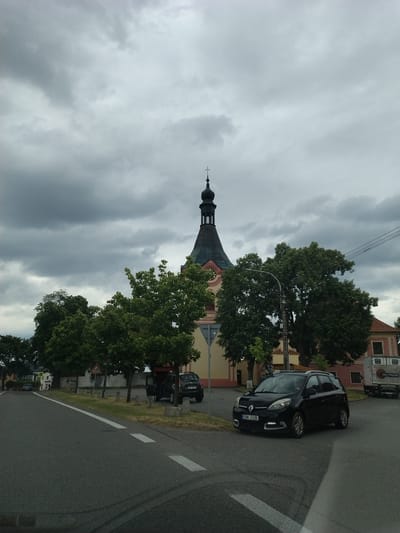 The image shows a street scene in Svatonice, Czechia, under a cloudy sky. A black Renault Scenic is parked on the right side of the road, facing left. To the left of the car, a church with a tall, dark steeple is visible, partially obscured by trees. In the background, there are buildings and more trees. A white truck with "VIN-TRANS" written on it is parked behind the Renault. The road has white lane markings.