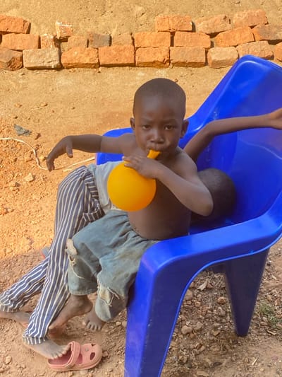 The scene, located outdoors in the city of Nev, Nigeria, depicts three children interacting with a blue plastic chair and an orange balloon. A young child, gender indeterminate, with dark skin, is seated front-center, shirtless, wearing torn light blue denim jeans. This child is actively blowing into an orange spherical balloon held to their mouth, looking directly forward. Partially visible behind this child and also in the blue chair are two other children. The second child's legs, clothed in blue and white striped pants, extend to the left foreground, with bare feet resting on reddish-brown dirt. One bare foot is positioned on a single pink Croc-style shoe. The third child's bare arm is visible draped over the far right side of the blue chair's backrest. The ground is unpaved, consisting of dry, reddish-brown earth mixed with small stones and debris. A coil of white electrical wire is present on the dirt to the left. The background features a low, weathered wall constructed from haphazardly stacked reddish-brown bricks. The scene is illuminated by strong natural sunlight.