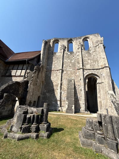 The image shows the exterior of the Walkenried Abbey in Walkenried, Germany. The structure is partially ruined, with large stone walls and arched windows visible. In the foreground, there are several large stone fragments and pillars on a grassy area. To the left, a section of the abbey is intact, featuring a red tiled roof and timber-framed walls. The sky is clear and blue.