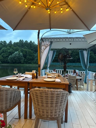 The image depicts an outdoor seating area at a restaurant located on the water in Zhukovka, Russia. The scene is set in the evening, as indicated by the warm glow of string lights overhead and the twilight sky.

In the foreground, two wooden tables are set for dining. The table on the left has two place settings with white plates, cutlery, and napkins. A glass of white wine and a glass of water are also present. A salt and pepper grinder and a small lamp sit in the center of the table. The table on the right also has a place setting. Two wicker chairs are placed in front of the tables.

In the background, on a raised deck, a large industrial fan is positioned on a stand. Two people are seated at a round table under a canopy, which is adorned with blue and white drapes. One person, a man with short, light-colored hair, is seated facing away from the camera. The other person, a woman with long, dark hair, is facing the camera. They appear to be engaged in conversation.

Beyond the deck, a body of water is visible, with a dense forest on the opposite bank. The trees are lush and green, suggesting it is summer or early autumn. The overall atmosphere is relaxed and inviting, with the ambiance of a waterfront dining experience.