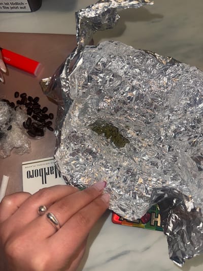 The image depicts an indoor scene in Rueil-Malmaison, France, viewed from an overhead perspective, displaying various items arranged on a light-colored, potentially marble or patterned, table surface. Central to the frame is a large, crumpled piece of aluminum foil, partially unfolded to reveal a quantity of green, leafy plant material, consistent with cannabis.
To the left of the foil, a small pile of dark, roasted coffee beans rests beside a clear, crinkled plastic material. Above these items, a red, cylindrical disposable vape pen is visible. In the lower left, an inverted white and red Marlboro cigarette pack is positioned, with a single, rolled white paper, possibly a cigarette or joint paper, adjacent to it.
A human hand, exhibiting manicured fingernails with French tips and a purple accent, occupies the lower foreground. This hand wears a silver-colored, open-style ring featuring a spherical and a teardrop-shaped element on the ring finger. The hand rests near the edge of the aluminum foil, partially obscuring a multi-colored object, possibly a tray or grinder, beneath the foil. In the upper left background, a dark object with partially visible white text, reading "jetzt auf," is present. The scene is illuminated by ambient or artificial light.