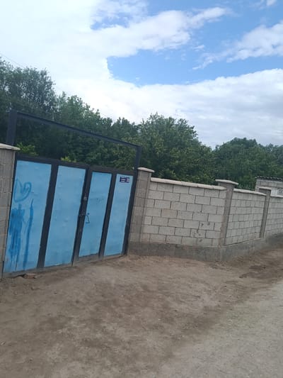 In Altinkul, Uzbekistan, a light blue metal gate with a black frame stands in front of a cinder block wall. The gate is divided into four vertical panels, with blue paint applied unevenly, showing vertical streaks and some rough markings. A padlock and a handle are visible on the gate. A small, white sign with blue text and the number "32" is affixed to the top right panel of the gate. The cinder block wall is constructed with grey blocks and has concrete pillars at intervals. The ground in front of the gate is dirt. Behind the gate and wall, a dense line of green trees is visible against a sky with scattered white clouds.