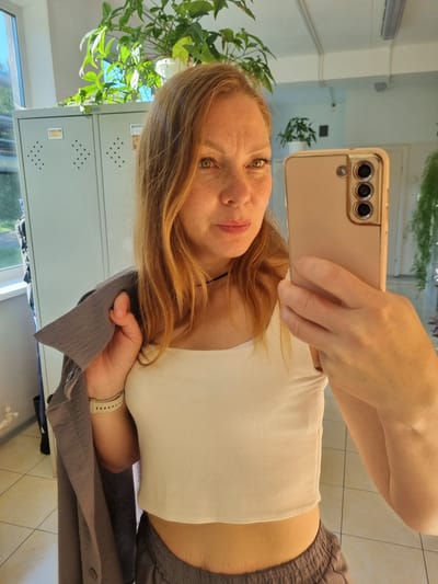 A blonde woman with freckles and light brown hair takes a selfie in a room. She is wearing a white ribbed crop top, grey sweatpants, and a grey jacket draped over her left shoulder. A smartwatch is visible on her left wrist. She is holding a rose-gold smartphone in her right hand, and the phone's camera is pointed at her reflection. The background shows metal lockers, a window, and plants. The floor is tiled. The scene appears to be indoors, possibly in a locker room or a similar facility. There is no indication of the location being in the city of Kemul, Russia.