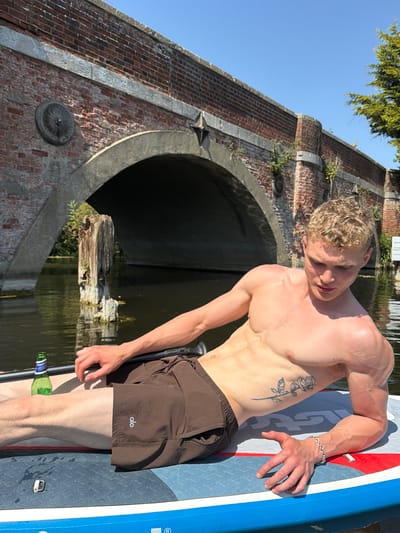 A shirtless, muscular man stands on a paddleboard in a canal in Babergh, United Kingdom. He wears brown shorts with the word "Alo" printed on them. A floral tattoo is visible on his left side. He has short, blond hair and a silver bracelet on his right wrist. A paddle rests beside him on the board, near a green glass bottle. Behind him, a large brick bridge spans the canal. The water is calm, and sunlight illuminates the scene.