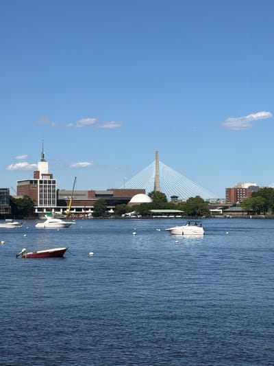 The image depicts a waterfront scene in Cambridge, Massachusetts, with the Museum of Science and the Zakim Bridge visible in the background. Several boats are scattered across the blue water, including a red dinghy in the foreground, a white speedboat further out, and other vessels anchored or moving on the water. The Museum of Science, a prominent brick building with a tall white spire, stands on the left side of the image. The Zakim Bridge, a distinctive cable-stayed bridge, dominates the skyline on the right. The sky is a clear blue with a few scattered white clouds. Trees and other buildings line the waterfront, suggesting an urban setting. The water is calm with gentle ripples, reflecting the bright sky. The scene conveys a peaceful, sunny day by the Charles River in Cambridge.
