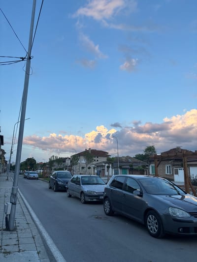 The image captures a street scene in Popovo, Bulgaria, under a blue sky with scattered cumulus clouds, some illuminated with golden light, indicating late afternoon or early evening. The perspective is from the sidewalk on the left, looking down the street.

In the foreground on the left, a tall, grey utility pole stands prominently, secured with black electrical wires connecting to various points. A small, light grey electrical box is attached to the base of this pole. The sidewalk, constructed from concrete slabs, shows some minor cracks and sparse patches of weeds. The road is asphalt, marked by a white painted curb separating it from the sidewalk.

Several vehicles are parked along the right side of the street. Closest to the viewer is a dark grey Volkswagen Golf hatchback, oriented with its front facing away from the camera. Behind it, a silver Peugeot 306 hatchback is partially visible. Further down the street, also on the right, is a dark grey SEAT Leon hatchback, and beyond that, another silver Peugeot hatchback.

Buildings line both sides of the street. On the right, various residential or mixed-use structures with tiled roofs are visible. One notable building features a light blue/turquoise door and a brick facade, while an adjacent structure has a prominent wooden pergola extending outwards. Another building further down has a white door. On the left side of the street, buildings are also visible, some with multiple windows and older architectural styles. Streetlights are interspersed along the street. Trees and vegetation are present between and behind the buildings, with some individual trees visible along the street. No discernible individuals are clearly visible in the foreground or midground.
