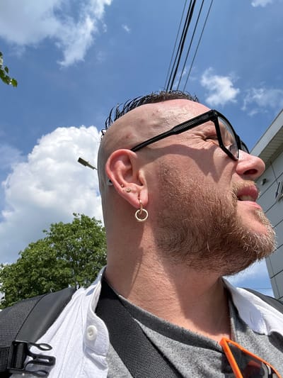 The image is a close-up, low-angle shot of a person with a shaved head, a beard, and glasses, taken outdoors in Selyatino, Russia. The person is looking upwards and to the right, with their mouth slightly open. They are wearing a grey t-shirt, a white collared shirt, and a backpack with black straps. There are visible ear piercings, including a hoop earring. The background shows a blue sky with white clouds, overhead power lines, and the top of a green tree. A street light is also visible in the distance. The lighting suggests it is daytime and sunny.