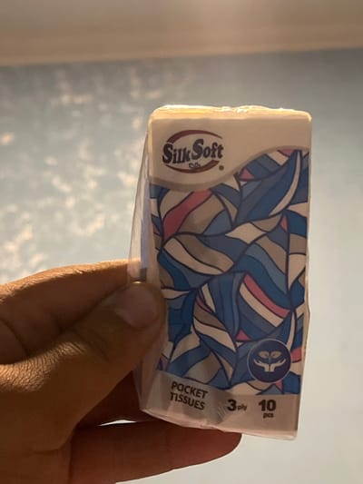 A close-up shot shows a hand holding a small, rectangular package of Silk Soft pocket tissues. The package features a cream-colored top with the brand logo and a colorful, abstract pattern of blue, pink, and grey wavy lines on the front. Below the pattern, the text "POCKET TISSUES 3 ply 10 pcs" is visible, along with a circular icon of two hands cradling a sprout. The background is a light blue wall with a textured pattern that resembles clouds. The lighting is warm, suggesting an indoor setting. There is no visible indication of the location being Jrarbi, Armenia.