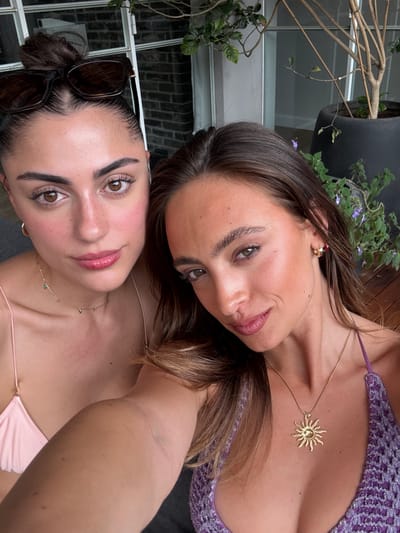 This is a close-up, tilted selfie featuring two young women, likely friends, in an outdoor setting, possibly a balcony or patio in Hod HaSharon, Israel. Both women have soft smiles, conveying contentment and friendliness under bright, natural daylight.
The woman positioned at the top right has medium-length brown hair and wears a purple crocheted top with thin straps. She is accessorized with a gold necklace featuring a distinct sun-shaped pendant and a small gold hoop earring. Her makeup is natural, with subtle eye definition and lip color.
The second woman, at the bottom left, has darker brown hair pulled back into a bun, with dark sunglasses resting on her head. She is wearing a light pink bikini top with thin straps and a delicate gold necklace interspersed with small green or blue stones. Her complexion shows a rosy blush on her cheeks.
The background includes various outdoor elements: green potted plants, bare tree branches, part of a wooden deck, and a window with a brick wall visible through it, suggesting an urban or suburban outdoor residential area. There is no visible text in the image.