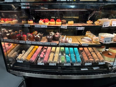 This image depicts the interior of a refrigerated display case filled with various desserts, located within a food retail establishment in McAllen, United States of America. The case contains three visible shelves populated with baked goods arranged for sale.
The top shelf features numerous small, round cheesecakes and tarts. Many are topped with fresh strawberries, raspberries, and blueberries. Price tags of approximately $5.49 are visible for individual items. Other larger, undecorated cheesecakes or tarts are also present.
The middle shelf primarily displays individual cupcakes and pastries. Multiple cupcakes in various flavors, including "Birthday Cake Cupcakes," "Holiday Cake Cupcakes," and "Strawberry Cupcakes," are arranged in paper liners. Prices range from $3.99 to $4.49 per cupcake. Additionally, two cream puffs and a chocolate-glazed eclair are visible on a wooden serving board, alongside smaller individual desserts in cups. A round cake with white frosting and colorful sprinkles, priced at $28.00, occupies the right side of this shelf.
The bottom shelf is predominantly filled with macarons of diverse colors, including blue, green, pink, brown, yellow, and off-white, presented on clear acrylic trays. Labels such as "Pistachio Macarons," "Dark Chocolate Macarons," "Salted Caramel Macarons," "Almond Macarons," and "Earl Grey Macarons" are displayed with prices around $1.99-$2.00 each. The far right section of this shelf contains several large, round multi-layered cakes, possibly tiramisu, with smooth brown tops and white sides, some featuring decorative elements like a single coffee bean or cocoa powder dusting.
The display case is illuminated, and a reflection of the store's ceiling lights is visible on the glass. No individuals are present within the frame. A "B10" label is affixed to the top interior of the case.