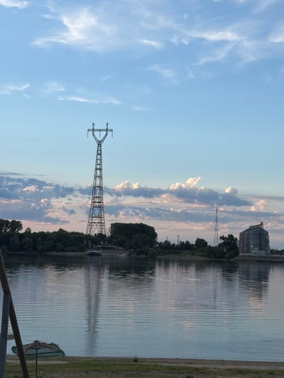 This is a wide-angle, eye-level, outdoor shot of a riverfront scene in Oryahovo, Bulgaria. The image captures a serene twilight atmosphere with a calm, reflective river in the foreground. On the far bank, a dense line of trees separates the water from industrial structures and a prominent high-voltage electricity pylon. To the right, a large, grey grain silo complex is visible. The sky above is a pale blue, adorned with wispy white and pink-tinged clouds. The reflection of the electricity pylon is clearly visible on the rippling surface of the river. In the immediate foreground, a small patch of sandy ground with scattered green grass is visible, along with a fragment of a wooden structure on the left and a partially visible beach umbrella structure. No people are present in the image.