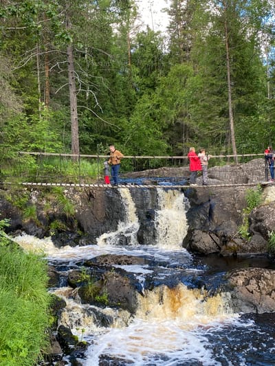 A narrow, suspension footbridge spans a rocky riverbed with a series of small waterfalls in Ruskeala, Russia. The bridge is constructed of wooden planks and rope, with several individuals walking across it. On the left side of the bridge, a man in a tan jacket and jeans stands with a young child wearing a red hat and red pants. Further to the right, a woman in a red jacket and a child in a gray hoodie are visible. On the far right, another individual in a dark jacket and jeans stands. The river below the bridge cascades over dark, wet rocks, creating white foam and turbulent water. The surrounding environment is a lush, green forest with tall trees and undergrowth. The lighting suggests it is daytime, with sunlight filtering through the trees.