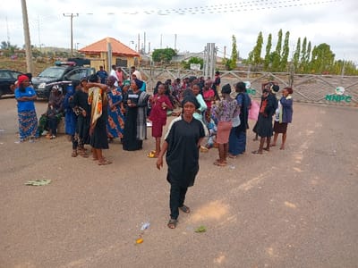 A group of mostly women are gathered in a paved area in front of a gated compound. The gate is adorned with the inscription “NNPC”. There is a building with a red roof in the background, which appears to be a guardhouse, and tall trees beyond the gate. The women appear to be engaged in conversation, with some sitting on the ground. A few men are also present. There is a dark-colored car parked to the left of the image, and another car, possibly a police vehicle, is parked on the left side of the frame. There are plastic water bottles and other debris scattered on the ground. The scene appears to be located in front of a facility owned by the Nigerian National Petroleum Corporation (NNPC) in the area of Ungwan Tanko, Nigeria.