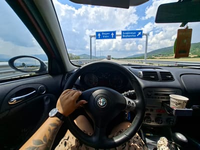 This first-person perspective image captures a person driving on a highway under a partly cloudy sky. The driver's hands, adorned with a watch and rings, are visible on the steering wheel of an Alfa Romeo. The dashboard and car interior are clearly in view, with a cup of coffee in a holder. In the background, a truck is visible on the road ahead, and rolling green hills frame the scene. Two blue road signs are prominently displayed, indicating directions to "Vidin" and "Bozhenitsa" in both Bulgarian and English. The time of day appears to be daytime, judging by the bright sunlight and the blue sky with scattered clouds. The overall impression is that of a road trip or commute through a rural or semi-rural landscape.