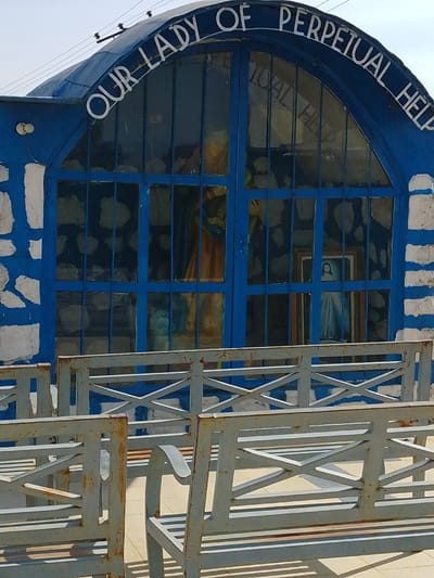 This image depicts the exterior of a religious shrine or chapel, identified by the inscription "OUR LADY OF PERPETUAL HELP," located in Bahit, Nigeria. The building features a prominent arched facade, painted a vibrant blue, with the lower walls exhibiting an irregular pattern of white stone or stucco interspersed with the blue. The arch is filled with multiple rectangular glass panels framed in blue, forming a grid.
Through these glass panels, internal features are visible, including a large, light-colored statue or figure positioned centrally, appearing to wear flowing, light-brown or gold-colored garments. To the right, a smaller framed image or statue depicting a figure in a light blue garment, characteristic of the Virgin Mary, is discernible. The interior background visible through the glass appears dark with some lighter, irregular shapes.
In the foreground, three rows of identical metal benches are arranged, facing the shrine. These benches are painted a light grey, and several display areas of rust, particularly along their edges and structural supports. Each bench features a slatted seat and backrest, with distinctive 'X'-shaped cross-braces on the side supports. The ground beneath the benches is a flat, light-colored surface, possibly concrete or tile. The sky above the structure is clear and light blue. No individuals are present in the scene.