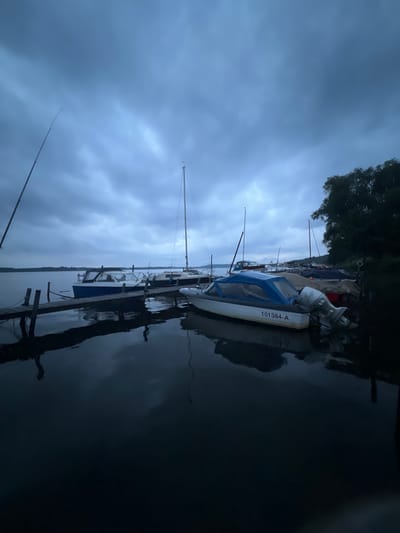 A gloomy, overcast day at the Schwielowsee in Germany, with a row of boats moored at a wooden jetty. The water is calm and dark, reflecting the cloudy sky and the boats. A few sailboats and motorboats are visible, some with covers on. The trees on the right bank of the lake are dark and silhouetted against the sky. The overall mood is somber and still, hinting at an approaching storm or the end of a day.